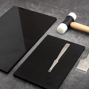May include: Two black cutting mats, a white and black rubber mallet, a metal stitching tool, and a metal awl. The tools are laid out on a gray surface.