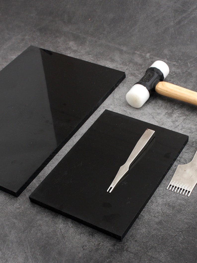 May include: Two black cutting mats, a white and black rubber mallet, a metal stitching tool, and a metal awl. The tools are laid out on a gray surface.