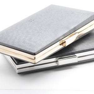 Puede incluir: Dos bolsos de mano rectangulares apilados uno encima del otro. El bolso superior tiene una superficie gris texturizada con ribete y cierre dorados. El bolso inferior tiene un diseño similar con ribete plateado.