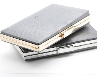 Può includere: Due pochette rettangolari sovrapposte. La pochette superiore ha una superficie grigia strutturata con finiture e chiusura color oro. La pochette inferiore ha un design simile con finiture color argento.