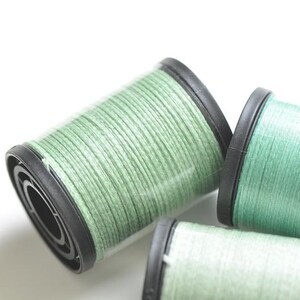 May include: Close-up of three spools of green thread. The thread is tightly wound around the spools, which have black plastic rims. The spools are in varying sizes and are set against a white background.