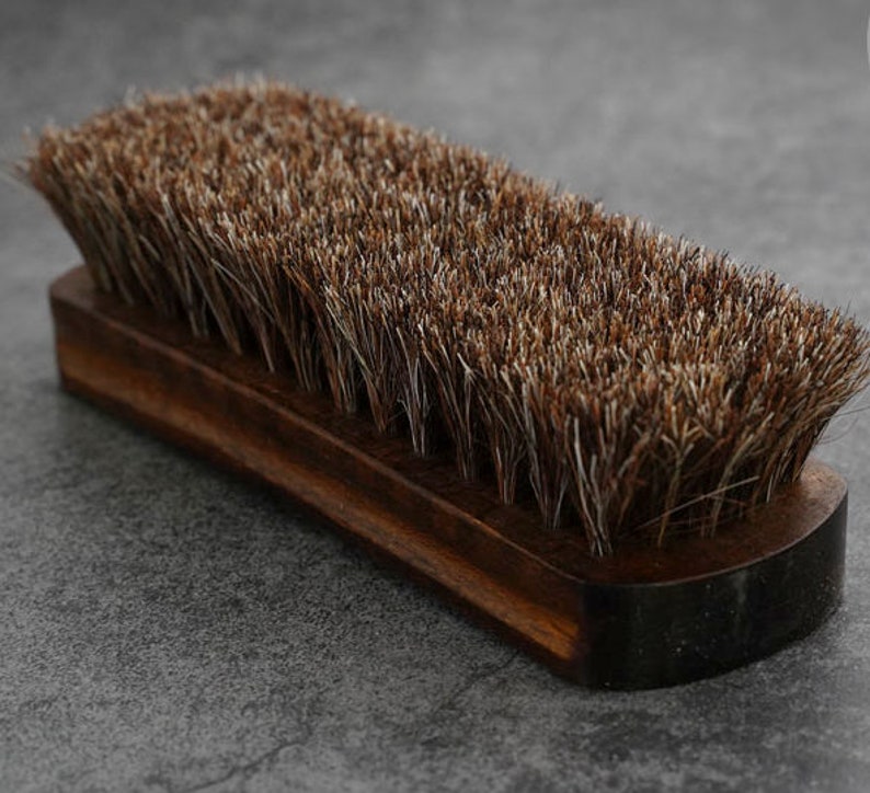 K&ouml;nnte beinhalten: Eine braune Holzschuhb&uuml;rste mit einer langen, dichten Borstenb&uuml;rste. Die B&uuml;rste ist zum Reinigen und Polieren von Lederschuhen gedacht.