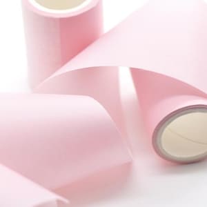 Könnte beinhalten: Nahaufnahme von drei Rollen rosa Papierband, von denen eine teilweise abgerollt ist. Das Band hat eine weiche, pastellrosa Farbe und die Rollen haben eine weiße Innenseite. Der Hintergrund ist weiß.