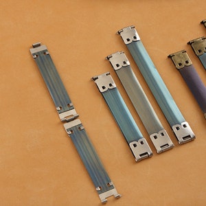 May include: Five metal strap buckles in various shades of blue and purple. The buckles are made of metal and have a brushed finish. The buckles are all the same size and shape, but they have different colors.