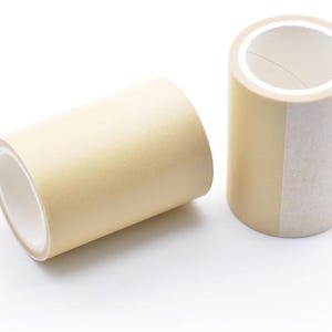 Könnte beinhalten: Zwei Rollen Abdeckband. Eine Rolle liegt auf der Seite, die andere steht aufrecht. Beide Rollen sind beige mit weißem Innenraum. Das Klebeband ist für Bastelarbeiten, Malerei oder den allgemeinen Gebrauch bestimmt.