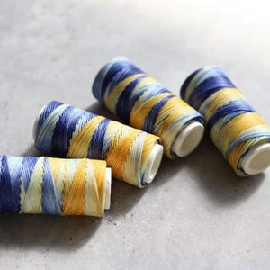 May include: Four spools of thread in shades of blue, yellow, and white. The thread is wound on white spools. The spools are arranged on a gray surface.