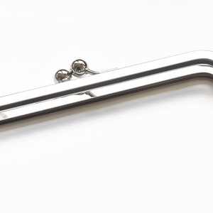 Puede incluir: Un marco de bolso plateado brillante, de forma rectangular con esquinas redondeadas. Presenta un diseño de doble barra y dos pequeños cierres esféricos. El metal es muy reflectante, sobre un fondo blanco. Ideal para marroquinería.