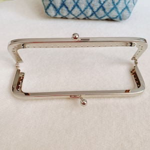 Op de afbeelding: Een zilverkleurig metalen tasframe met een rechthoekige vorm en afgeronde hoeken. Het frame heeft twee kleine, ronde, zilverkleurige metalen knoppen aan de boven- en onderkant.