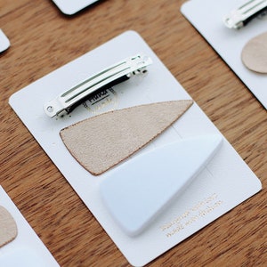 Peut inclure: Deux ensembles de pinces à cheveux dans un schéma de couleurs blanc et beige. Chaque ensemble comprend une petite pince métallique rectangulaire et une pièce décorative en forme de triangle. Les pièces décoratives sont faites d'un matériau doux et texturé. L'emballage est blanc avec des lettres dorées qui indiquent "Fabriqué avec amour".