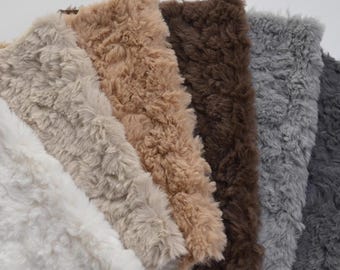 May include: Close-up of a collection of faux fur fabric swatches in various neutral colors. The swatches include white, beige, tan, brown, gray, and dark gray. The fabric has a soft, textured appearance.