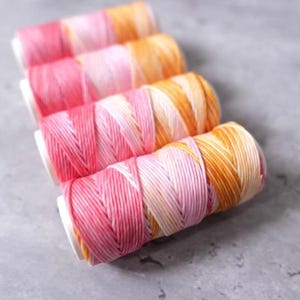 May include: Four spools of colorful thread in shades of pink, orange, and yellow. The thread is tightly wound on white spools, creating a gradient effect. Ideal for crafting and sewing projects.