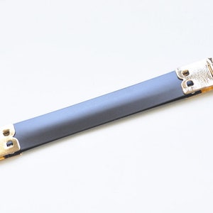 Könnte beinhalten: Ein graues Lederband mit goldenen Metallschließen an jedem Ende. Das Band ist etwa 25 cm lang.