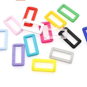 May include: A selection of brightly coloured rectangular plastic rings in shades such as red, pink, yellow, green, blue, purple, grey, black, and clear. The rings are scattered on a white surface.