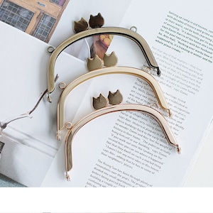 May include: Three gold-toned metal purse frames with cat-shaped clasps. The frames are stacked on top of each other, with the largest frame on the bottom and the smallest frame on top.