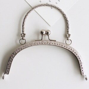 May include: Silver-toned metal purse frame with a decorative floral design. The frame has a rounded top and two loops on either side.