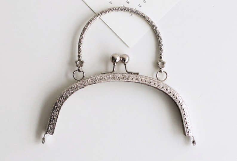 May include: Silver-toned metal purse frame with a decorative floral design. The frame has a rounded top and two loops on either side.