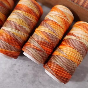 May include: Close-up of several spools of colorful waxed thread. The thread is wound around white spools and features a gradient of orange, yellow, and brown hues. The spools are arranged on a light gray surface.