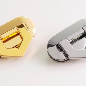 May include: Two metal purse clasps, one gold and one silver, are displayed on a white surface. Each clasp has a rectangular body with a geometric, angular design. The gold clasp is on the left, and the silver clasp is on the right.