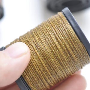 May include: Close-up of a spool of gold-colored thread. The thread is tightly wound around a black plastic core. The thread has a slight sheen, suggesting it may be metallic or have a reflective quality. The background is white.