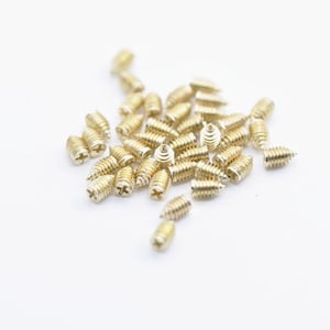 May include: A scattering of small, gold-coloured screws on a white surface. Each screw has a slotted head and a threaded body, designed for fastening materials together. The screws are likely made of brass or a similar metal.