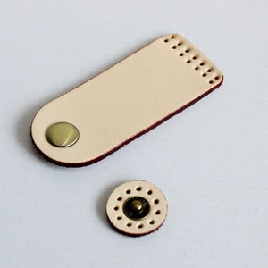 Puede incluir: Una correa de cuero de color crema con un borde cosido rojo y un botón de latón. La correa está diseñada para ser utilizada como hebilla o cierre para un bolso u otros artículos de cuero.