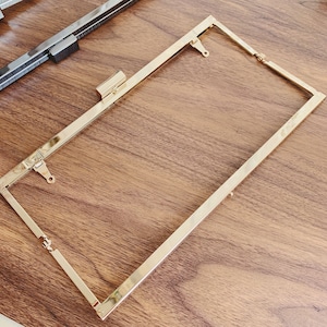 Puede incluir: Marco de metal dorado para un bolso de mano o una cartera. El marco es rectangular y tiene un cierre en un lado.