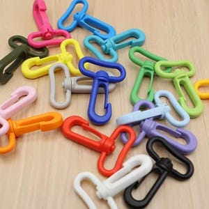 以下が含まれることがあります： ピンク、青、緑、黄色、オレンジ、赤、白、黒、紫など、さまざまな色合いのカラフルなスイベルスナップフックのコレクション。これらのプラスチックフックは、ストラップやその他のアイテムに取り付けるように設計されています。