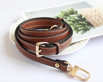 May include: A brown leather strap with a gold buckle and a gold hook clasp. The strap is folded and coiled on a white surface.