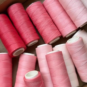 Può includere: Una collezione di rocchetti di filo rosa e rosso, con sfumature che vanno dal rosa chiaro al rosa intenso. I rocchetti sono avvolti strettamente, mostrando la trama del filo. L'immagine suggerisce un tema artigianale o di cucito.