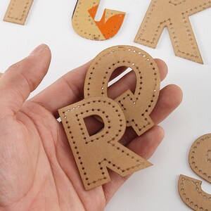 May include: Two brown cardboard letters, R and Q, with a dotted stitch design around the edges. The letters are held in a hand.