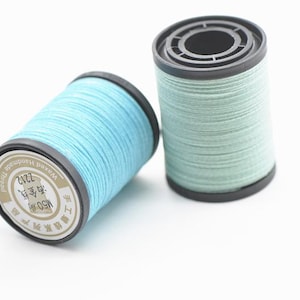 Puede incluir: Dos carretes de hilo encerado hecho a mano en tonos azules y verdes. Los carretes tienen bordes y bases negros. Un carrete está inclinado, revelando una etiqueta con caracteres chinos y el número 2212.