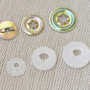 May include: Assortment of metal and plastic snap fasteners in various sizes. The metal fasteners have a star-shaped opening and a gold finish. The plastic fasteners are white and have a round opening.