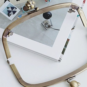 Puede incluir: Un marco de metal dorado para un bolso o un clutch. El marco está abierto y tiene un cierre en forma de pata.