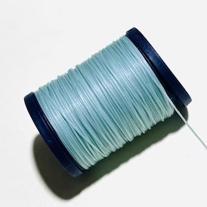 May include: A spool of light blue thread with a dark blue plastic core. The thread is tightly wound around the core, and a single strand extends from the spool. The background is white.
