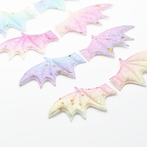 May include: A collection of glittery, pastel-colored bat wings in shades of pink, purple, blue, and yellow. Each wing has a sparkly finish and small gold star accents. The wings are arranged on a white surface.