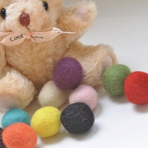 May include: A plush teddy bear with a "Love Love" ribbon sits on a white surface with a variety of colourful felt balls. The felt balls are in shades of purple, green, pink, cream, yellow, orange, blue, and black.