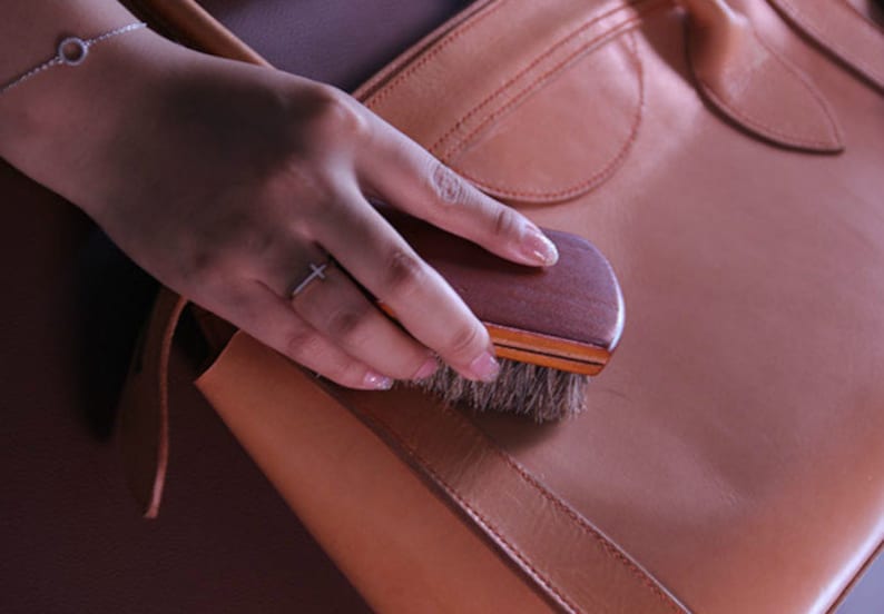 K&ouml;nnte beinhalten: Eine Hand h&auml;lt eine kleine Holzb&uuml;rste mit Naturborsten und b&uuml;rstet eine braune Ledertasche. Die Tasche hat einen Klappdeckel und eine gen&auml;hte Naht.