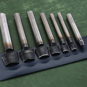 May include: Set of eight metal leather hole punches with different sizes. The punches are arranged in order of size, with the largest punch on the left and the smallest punch on the right. The punches are on a blue leather background.