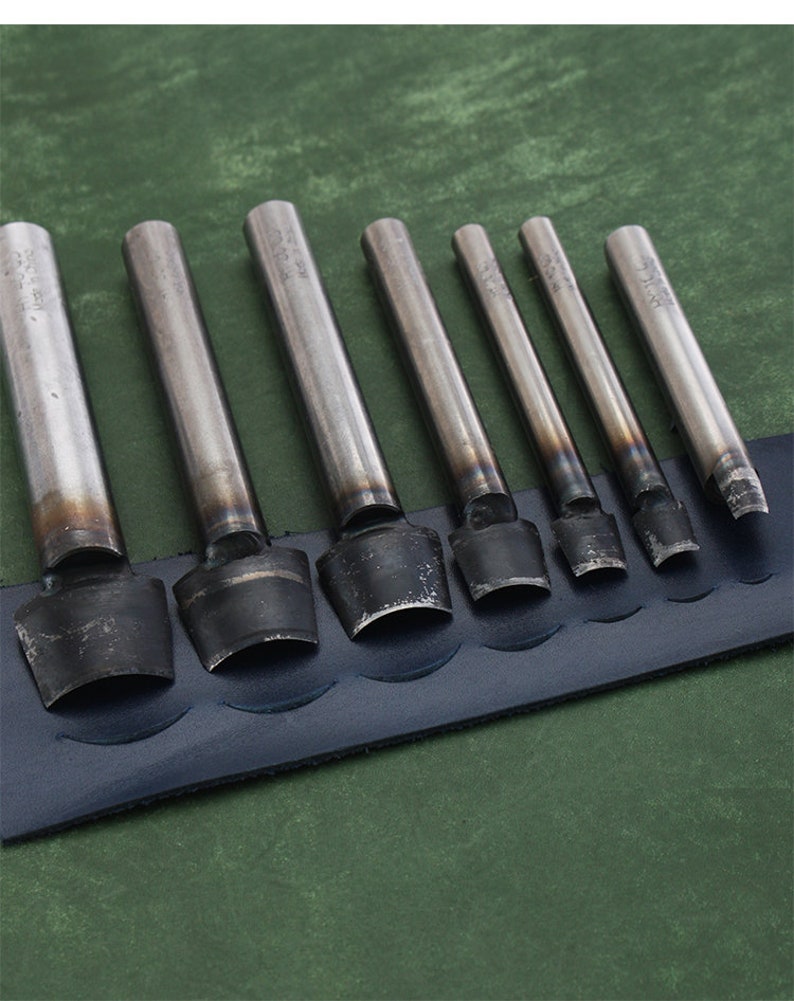 May include: Set of eight metal leather hole punches with different sizes. The punches are arranged in order of size, with the largest punch on the left and the smallest punch on the right. The punches are on a blue leather background.