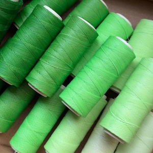 Puede incluir: Una colección de carretes de hilo verde en varios tonos, desde oscuros hasta claros, dispuestos juntos. Los carretes tienen núcleos blancos y están bien enrollados con hilo, creando una superficie texturizada. La imagen muestra una gama de tonos verdes.
