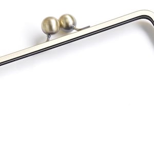 Pode incluir: Uma armação de metal para bolsa com formato retangular e cantos arredondados. A armação tem acabamento em latão e dois pequenos fechos redondos em forma de bola. A armação é projetada para ser costurada em uma bolsa.