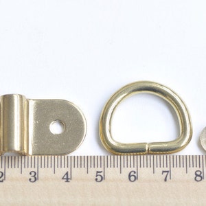 May include: Four gold-toned metal hardware pieces for a bag or other project. The pieces include a D-ring, a flat plate with two holes, and two rivets.