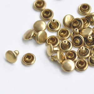 May include: A collection of small, brass-colored metal rivets scattered on a white surface. Each rivet has a rounded head and a post for securing to fabric or leather. The rivets are likely used for crafting or adding decorative elements.
