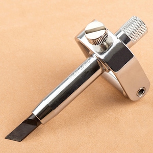 May include: A silver leather crafting tool with a black blade and a knob for adjusting the depth of the cut. The tool is labeled "CRAFT JAPAN".
