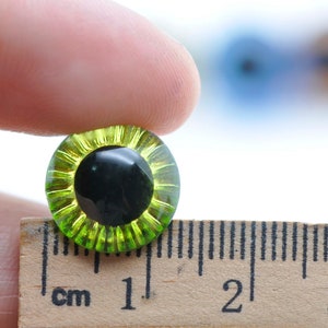 May include: A close-up of a green and black plastic safety eye, measuring 1 cm in diameter, for use in crafts and doll making.
