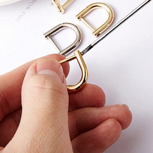 Puede incluir: Tres hebillas de anillo en D en acabados dorado, plateado y níquel. Las hebillas se muestran sobre una superficie blanca.