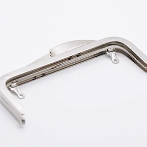 May include: A silver-colored metal purse frame with a curved, rectangular shape. The frame has a hinged closure and small loops on each end for attaching to a bag. The frame is designed for use in crafting.
