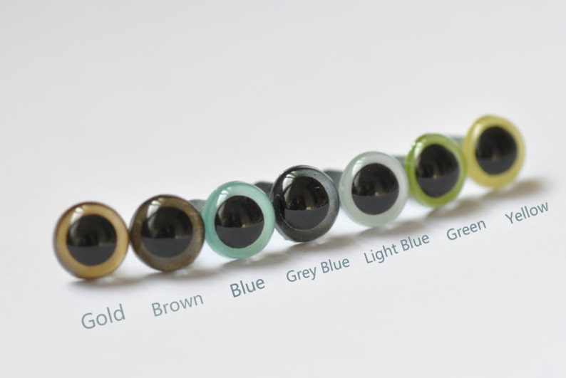 May include: A row of ten colorful plastic safety eyes for crafts, arranged from left to right: gold, brown, blue, grey blue, light blue, green, and yellow.