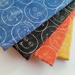 Op de afbeelding: Vier stofstalen met een herhalend patroon van witte lachende gezichten op verschillende gekleurde achtergronden. De stalen zijn op elkaar gestapeld, met de blauwe staal bovenop, gevolgd door zwart, oranje en geel.