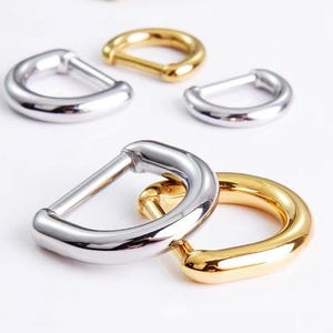 Puede incluir: Primer plano de varios herrajes de anilla en D en plata y oro. Las anillas en D son de metal y tienen un acabado liso y brillante. El herraje se utiliza probablemente para bolsos, cinturones u otros accesorios.